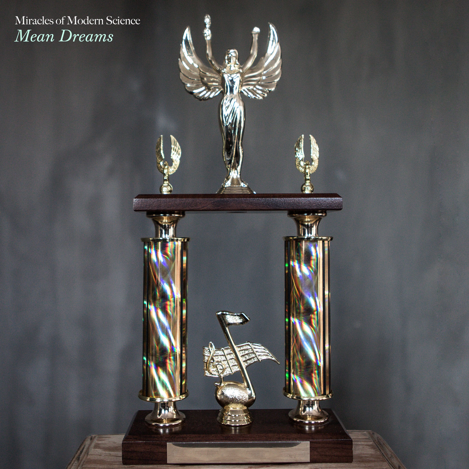 Miracles of Modern Science 'Mean Dreams' album cover: Ornate trophy with winged figure atop, colorful holographic columns, and musical note figurine on dark wooden base.